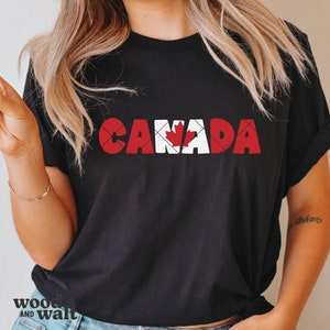 May include: Black t-shirt with the word "CANADA" in red and white block letters. The letter "A" features a red maple leaf design, a symbol of Canada. The shirt is a casual style.