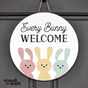 May include: A round white door hanger with the text "Every Bunny Welcome" in black. Three bunny silhouettes are below the text, one pink, one yellow, and one blue.