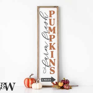 May include: A rectangular wooden sign with the words "Farm Fresh Pumpkins" in black and orange lettering. The sign is framed in a light brown wood. Decorative pumpkins and gourds are placed at the base of the sign. The sign also indicates "5 miles".