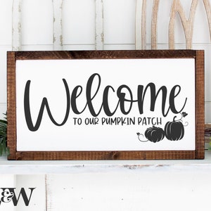 May include: A white wooden sign with a brown frame, featuring the text "Welcome to our pumpkin patch" in black script font. Two black silhouette pumpkins with vines are displayed below the text.