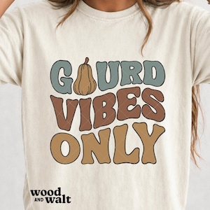 May include: A cream-colored short-sleeve t-shirt featuring the text GOURD VIBES ONLY in a retro font. The word GOURD is in muted blue with a brown pumpkin icon replacing the letter O. VIBES is in brown, and ONLY is in tan. Wood and Walt is printed below.