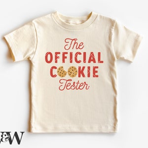 May include: A cream coloured t-shirt with red text that reads "The Official Cookie Tester" with two brown chocolate chip cookies on the shirt.