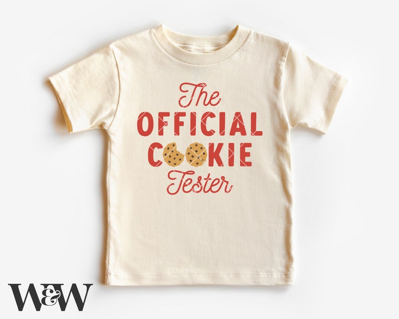 May include: A cream coloured t-shirt with red text that reads "The Official Cookie Tester" with two brown chocolate chip cookies on the shirt.