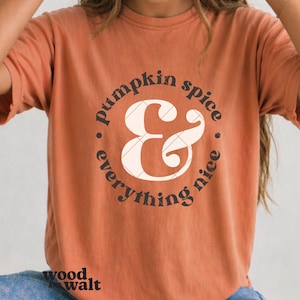 May include: A burnt orange short-sleeve t-shirt featuring a large white ampersand symbol with a subtle grid pattern at the centre. The text "pumpkin spice & everything nice" is printed in dark brown letters in a circular layout around the symbol.