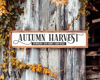 Könnte beinhalten: Ein rechteckiges Holzschild mit weißem Hintergrund und dunkelbraunem Rahmen. Der Text lautet AUTUMN HARVEST in großen schwarzen Buchstaben, wobei die Wörter PUMPKINS • HAY RIDES • CORN MAZE in kleinerer schwarzer Schrift auf einem Balken stehen.