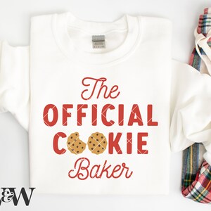 May include: White sweatshirt with red text that says "The Official Cookie Baker" with two chocolate chip cookies on the word "cookie".