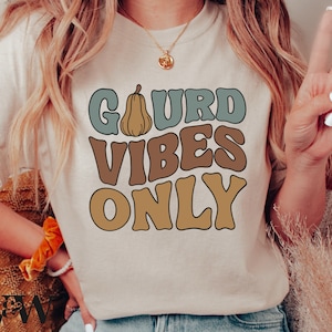 Op de afbeelding: Een beige T-shirt met de tekst "Gourd Vibes Only" in een retro-lettertype. De tekst is in tinten bruin, blauw en geel. Boven de tekst staat een kleine pompoenafbeelding.