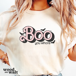 May include: Cream-colored t-shirt with the text "Boo you whore" in a retro font with pink and black accents. The design includes bats and stars, perfect for Halloween.