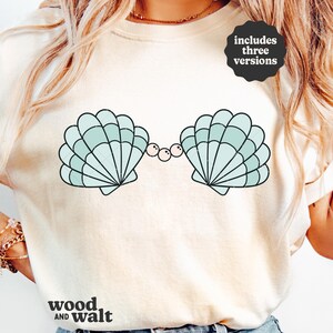 May include: Cream-colored sweatshirt featuring a light blue seashell design with black outlines and pearl accents. The design includes two shells and the text "includes three versions". The brand name "wood and walt" is printed at the bottom.