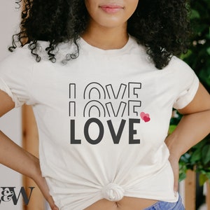 May include: A white t-shirt with the words "I LOVE LOVE LOVE" printed in black. The word "LOVE" is repeated three times, with the first two "LOVE" words in a stylized font with a rainbow arch design. A small pink heart is next to the last "LOVE" word.