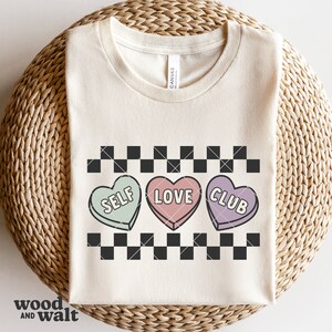 May include: A white t-shirt with a black and white checkered pattern, three candy hearts with the words "Self Love Club" on them.