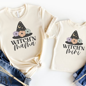 Può includere: Due magliette color crema con una grafica di cappello da strega e fiori. La maglietta più grande dice "Witch'n Mama" e quella più piccola dice "Witch'n Mini".