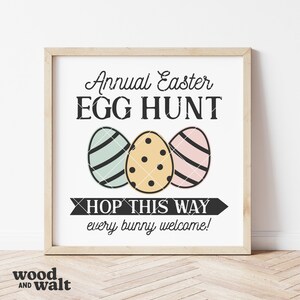 Op de afbeelding: Een wit bord met een houten frame dat een jaarlijkse paaseierenjacht aankondigt. Het bord toont drie kleurrijke paaseieren en de tekst "HOP THIS WAY every bunny welcome!"