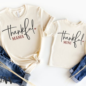 Puede incluir: Dos camisetas de color crema con el texto "thankful" en letra cursiva negra. La camiseta más grande tiene el texto "mama" en color marrón debajo de la palabra "thankful". La camiseta más pequeña tiene el texto "mini" en color marrón debajo de la palabra "thankful". Ambas camisetas tienen un pequeño corazón rojo a la derecha de la palabra "thankful".