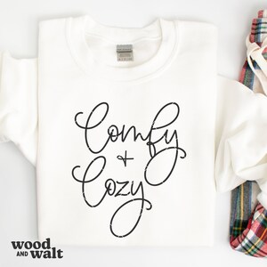 Op de afbeelding: Witte trui met zwarte tekst die "Comfy + Cozy" zegt.