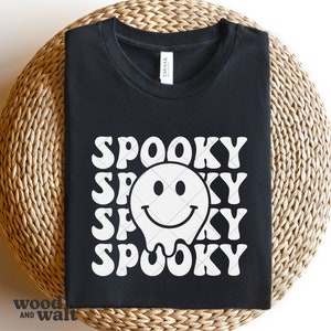 May include: A black t-shirt featuring a white graphic print of a melting smiley face surrounded by the repeating word SPOOKY in a wavy, bold font. The shirt has a crew neckline and a white brand label visible at the inside of the collar.