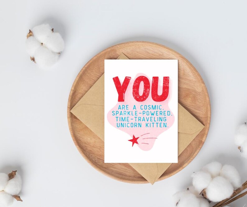 Puede incluir: Una tarjeta blanca con texto rosa y rojo que dice "YOU ARE A COSMIC. SPARKLE-POWERED. TIME-TRAVELING UNICORN KITTEN." Una nube rosa con una estrella roja y una estrella fugaz azul en el fondo.
