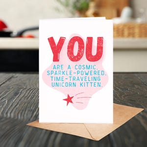 Puede incluir: Una tarjeta de felicitaci&oacute;n rosa y blanca con el texto "YOU ARE A COSMIC. SPARKLE-POWERED. TIME-TRAVELING UNICORN KITTEN." en letras azules y rojas.