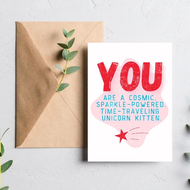 Puede incluir: Una tarjeta rosa y blanca con el texto "YOU ARE A COSMIC. SPARKLE-POWERED. TIME-TRAVELING UNICORN KITTEN." en letras rojas y azules. Una estrella roja con una cola rosa se encuentra en la esquina inferior derecha.