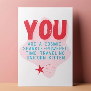 Puede incluir: Una tarjeta rosa y blanca con el texto "YOU ARE A COSMIC. SPARKLE-POWERED. TIME-TRAVELING UNICORN KITTEN." en letras azules y rojas. Una estrella roja con contorno blanco se encuentra en la esquina inferior derecha.