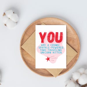 Puede incluir: Una tarjeta blanca con texto rosa y rojo que dice "YOU ARE A COSMIC. SPARKLE-POWERED. TIME-TRAVELING UNICORN KITTEN." Una nube rosa con una estrella roja y una estrella fugaz azul en el fondo.