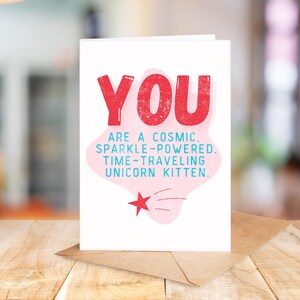 Puede incluir: Una tarjeta blanca con texto rosa y azul que dice "YOU ARE A COSMIC. SPARKLE-POWERED. TIME-TRAVELING UNICORN KITTEN." Hay una nube rosa con una estrella roja y una estrella fugaz azul.