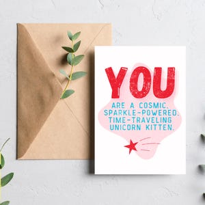 Puede incluir: Una tarjeta rosa y blanca con el texto "YOU ARE A COSMIC. SPARKLE-POWERED. TIME-TRAVELING UNICORN KITTEN." en letras rojas y azules. Una estrella roja con una cola rosa se encuentra en la esquina inferior derecha.