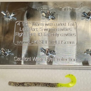 May include: A rectangular aluminum fishing lure mold with five wing nuts on the surface. Engraved text reads: 4 1/2" Worm with curled Tail, Left Part: 5 worm cavities, Right Part: 10 Tail only cavities, www.LAKESIDEMOLDS.com, Caution! Wear Eye Protection. A single 4.5-inch plastic worm lure with a yellow curled tail is placed in front.