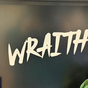 May include: The word "WRAITH" in a bold, stylized font is displayed in white against a dark background. The letters have sharp, angular edges, creating a striking visual contrast. The overall aesthetic suggests a modern, edgy design.