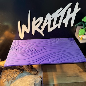 May include: A rectangular, purple, wood-grain textured surface. The word "WRAITH" is written in white, stylized lettering above the surface. A small decorative plant in a pot is on a shelf to the right.