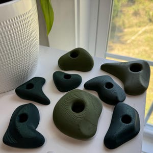 May include: A set of eight green and dark green climbing holds for a home climbing wall. The holds are made of a textured plastic and have different shapes and sizes.