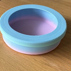 May include: A small, round, pastel-colored container with a light blue rim and a pink, blue, and green ombre finish. The container is made of wood and has a smooth, polished surface.
