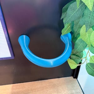 May include: A bright blue, U-shaped handle, possibly 3D-printed, is mounted on a black surface. The handle has a smooth, curved design with rounded ends. The background includes artificial green leaves and a wooden floor.