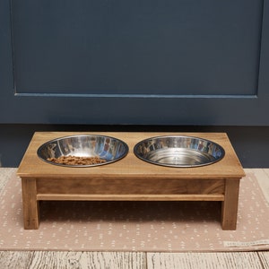 Puede incluir: Un soporte de madera elevado para cuencos de comida y agua para mascotas con dos cuencos de acero inoxidable. Un cuenco est&aacute; lleno de comida para mascotas de color marr&oacute;n. El soporte est&aacute; sobre una alfombra beige con estampado.