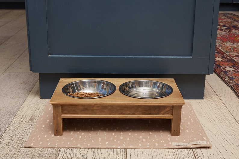 Puede incluir: Un soporte de madera elevado para cuencos de comida y agua para mascotas con dos cuencos de acero inoxidable. Un cuenco est&aacute; lleno de comida para mascotas de color marr&oacute;n. El soporte est&aacute; sobre una alfombra beige con estampado.