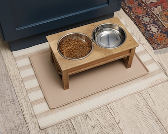 May include: A wooden elevated dog bowl stand with two stainless steel bowls. One bowl is filled with brown dog food. The stand is on top of a beige and white striped mat.