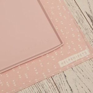 Puede incluir: Un mantel individual de silicona rosa con un borde elevado y el texto "Helden Pets" impreso en &eacute;l. El mantel individual est&aacute; sobre un mantel individual de tela rosa con un patr&oacute;n de corazones blancos.
