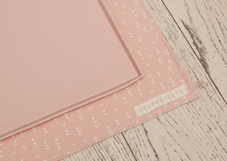 Puede incluir: Un mantel individual de silicona rosa con un borde elevado y el texto "Helden Pets" impreso en &eacute;l. El mantel individual est&aacute; sobre un mantel individual de tela rosa con un patr&oacute;n de corazones blancos.