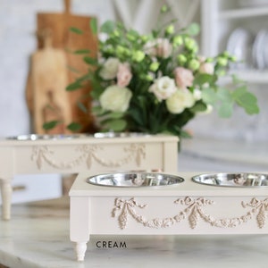Pu&ograve; includere: Due ciotole per cibo per animali domestici in ceramica color crema con bordi rialzati e motivi floreali decorativi. Le ciotole sono su un supporto abbinato con quattro gambe. Il supporto ha la scritta "CREAM" stampata sul davanti.