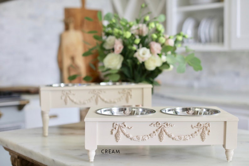 Pu&ograve; includere: Due ciotole per cibo per animali domestici in ceramica color crema con bordi rialzati e motivi floreali decorativi. Le ciotole sono su un supporto abbinato con quattro gambe. Il supporto ha la scritta "CREAM" stampata sul davanti.