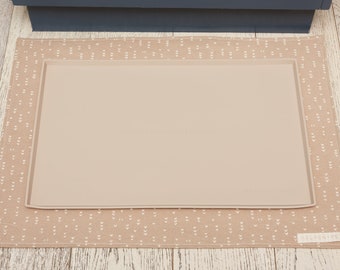 May include: A beige silicone mat with a white patterned fabric border. The mat is placed on a light wood floor. The fabric border has the text "HELFN + DEIN" printed on it.
