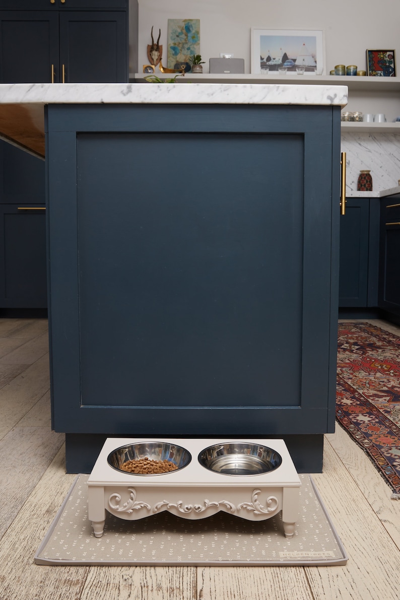 Puede incluir: Un soporte de cuenco para comida de mascotas de cer&aacute;mica blanca con dos cuencos de acero inoxidable. El soporte tiene detalles ornamentados y se encuentra sobre una alfombra gris con estampado delante de un armario azul oscuro.