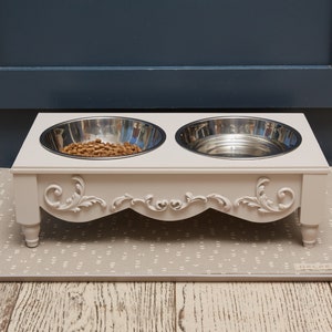 Puede incluir: Un soporte elevado blanco para cuencos de comida y agua para mascotas con dos cuencos de acero inoxidable. El soporte tiene detalles ornamentales y se encuentra sobre una alfombra gris con estampado.