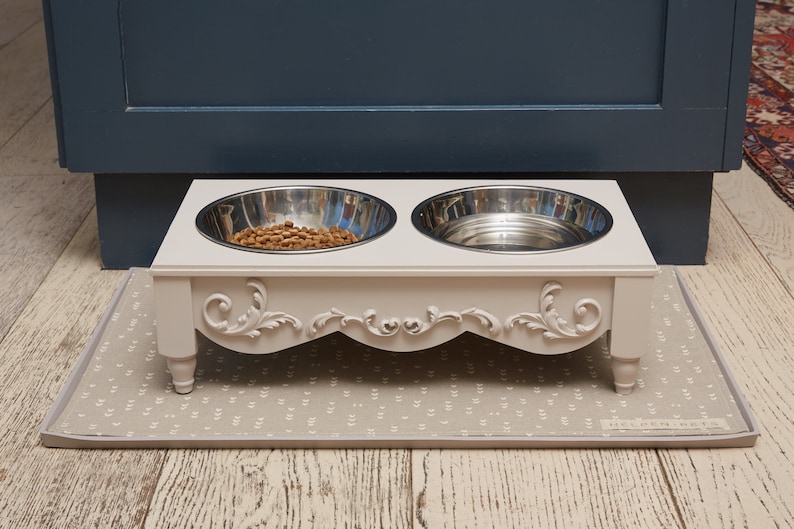Puede incluir: Un soporte elevado blanco para cuencos de comida y agua para mascotas con dos cuencos de acero inoxidable. El soporte tiene detalles ornamentales y se encuentra sobre una alfombra gris con estampado.