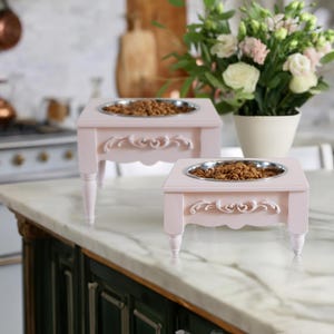 May include: Two pink elevated pet food bowls with ornate detailing and raised legs. The bowls are filled with dry pet food and are on a white countertop.