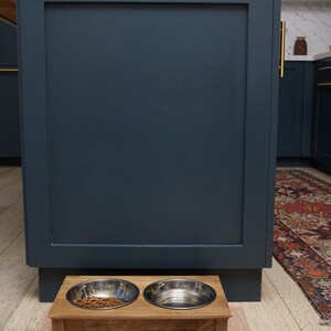 Puede incluir: Un soporte de madera para comida de mascotas con dos cuencos de acero inoxidable. Un cuenco est&aacute; lleno de comida de mascotas marr&oacute;n. El soporte est&aacute; sobre una alfombra de color marr&oacute;n claro con estampado.
