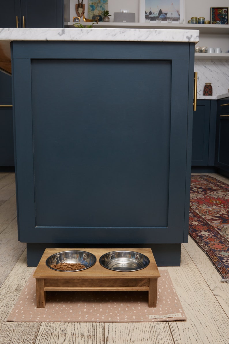 Puede incluir: Un soporte de madera para comida de mascotas con dos cuencos de acero inoxidable. Un cuenco est&aacute; lleno de comida de mascotas marr&oacute;n. El soporte est&aacute; sobre una alfombra de color marr&oacute;n claro con estampado.