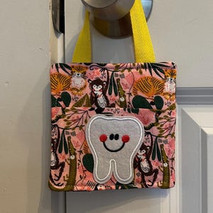 May include: A small, square fabric tooth fairy bag with a yellow handle hangs from a doorknob. The bag features a pink background with a jungle-themed pattern and a smiling tooth applique.