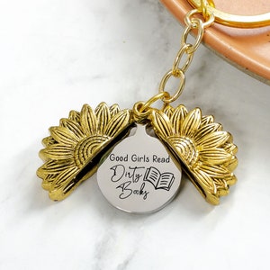 May include: A gold-colored sunflower keychain with a round pendant that reads "Good Girls Read Dirty Books" and a small book illustration. The keychain has a gold ring and chain.