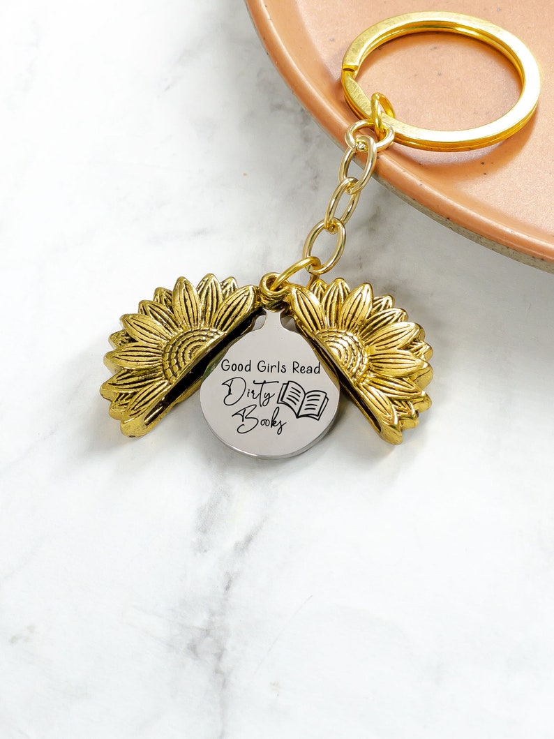 May include: A gold-colored sunflower keychain with a round pendant that reads "Good Girls Read Dirty Books" and a small book illustration. The keychain has a gold ring and chain.